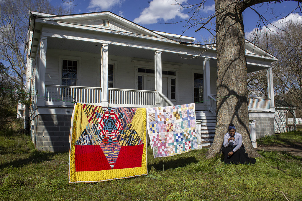 “All in a Day’s Work pt. 1,” by Amber Quinn. Black woman dressed as a slave crouching by tree smoking a cigarette, quilts are strung across a clothes line to her left.