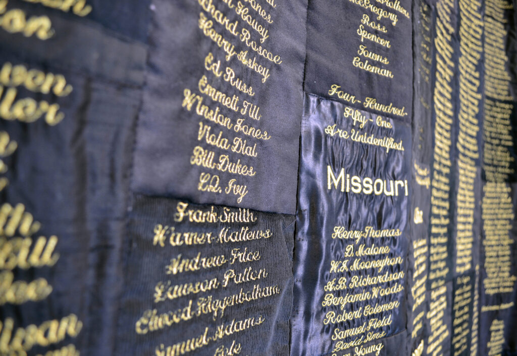 Detail shot of "Strange Fruit" by April Shipp, Just Injust 2021. Close-up of names of lynching victims embroidered in gold to quilt of patches of black fabric.